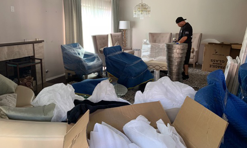 Mover in a living room arranging metal stools amid blue moving blankets, packed boxes, and wrapped furniture pieces.