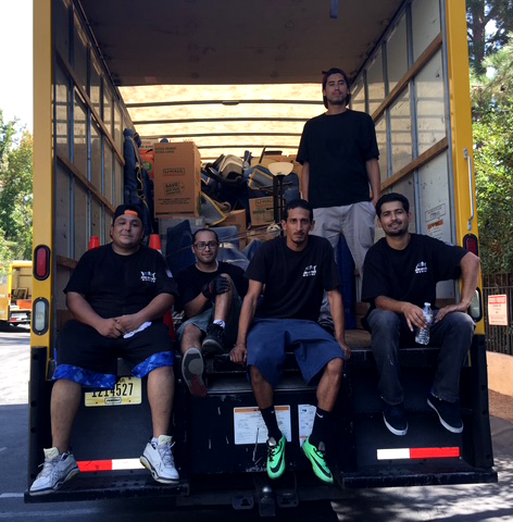 Five men sit and stand in the open back of a yellow moving truck, surrounded by packed boxes and furniture.