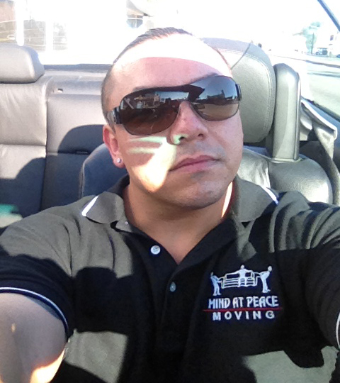 Man wearing a black polo with the Mind At Peace Moving logo sits in a car wearing dark sunglasses.